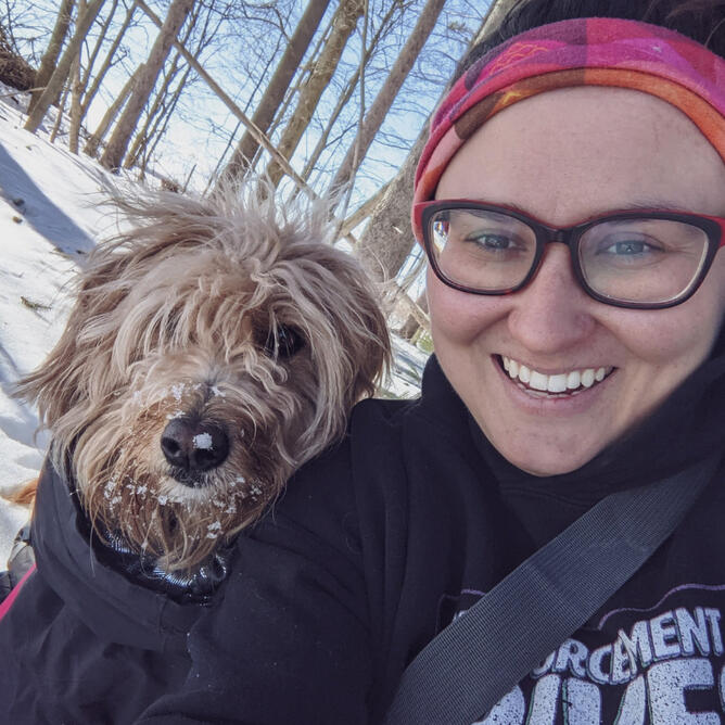 Klarissa is a femme non-binary person with brown hair, Chelsea is a blonde mini doodle, they are in a snowy forest.