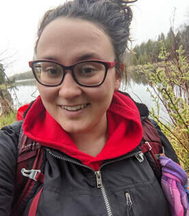 Klarissa I am a white, femme non-binary person with brown hair & brown eyes. I wear black, plastic framed glasses & hair pulled into a messy bun. I am wearing a red sweater, black jacket, with a red backpack on the shore of a pond.