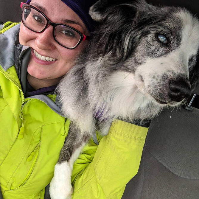 Klarissa is a femme non-binary person with brown hair & glasses. They are wearing a lime green rain coat, while Ellie, a blue merle mini Aussie stands over their shoulder.